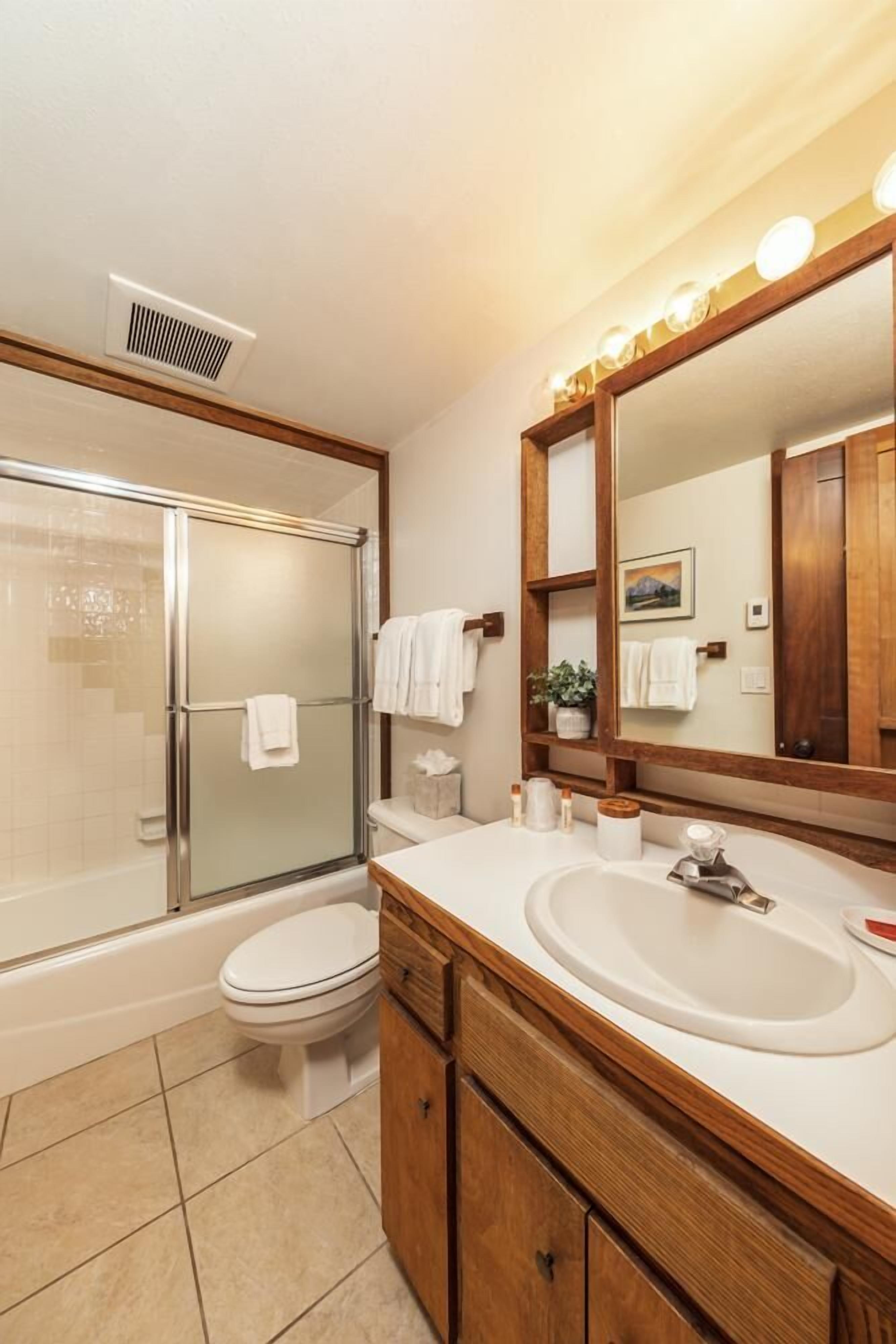 Une salle de bain moderne avec une douche à parois de verre, des toilettes blanches, un meuble-lavabo en bois et un grand miroir avec des lumières au-dessus.