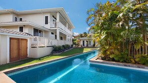 Una casa de dos pisos con piscina, rodeada de abundante vegetación y palmeras.