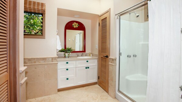 Un baño con cabina de ducha de vidrio, tocador con espejo y ventana con persianas.