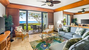 A spacious living room with a large glass door leading to a balcony with a view of the ocean. There are wicker chairs, a sofa, and a coffee table with a floral rug. - Poipu Kapili 55 By Parrish Kauai- fabulous oceanfront & sunset views (Koloa)
