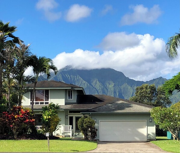 A two-story house with a garage, surrounded by lush greenery and a mountain backdrop.