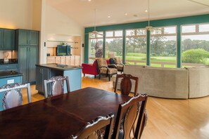 A modern kitchen with dark cabinetry, a dining area with a wooden table and chairs, and a large window offering a view of a green landscape. - Modern AC Home w/ Golf Course & Mountain Views! (Princeville)