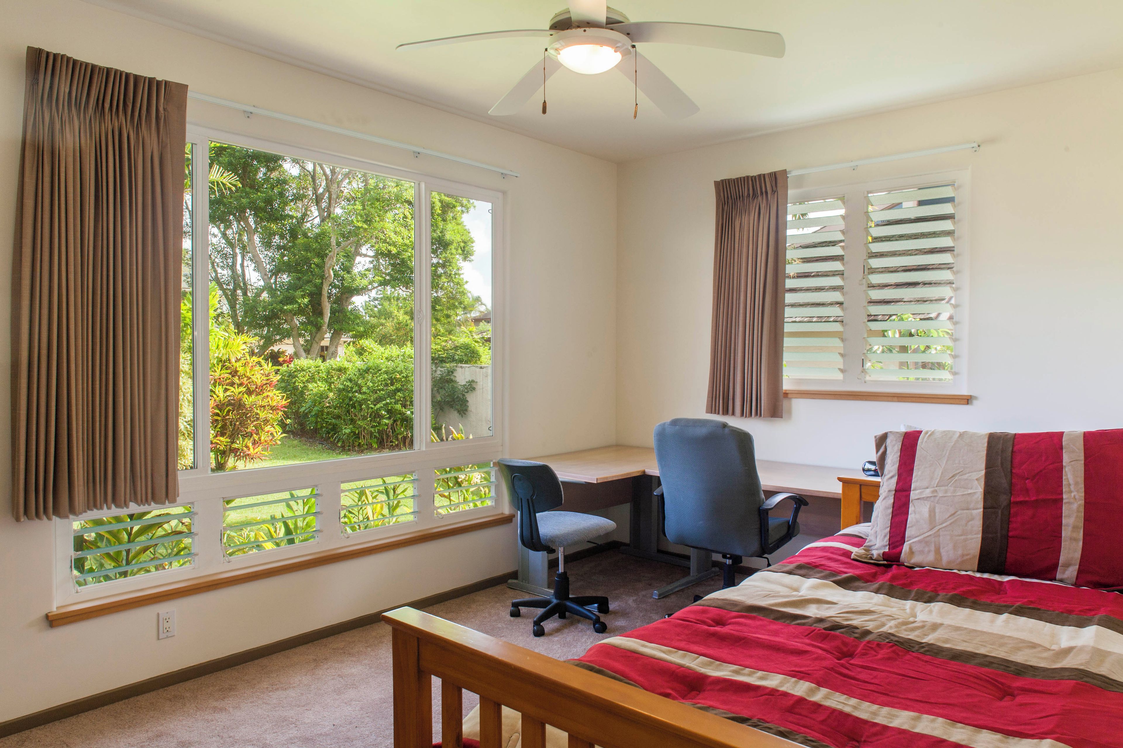 Une chambre à coucher comprenant un lit, un bureau et une chaise, de grandes fenêtres avec des rideaux et un ventilateur de plafond.