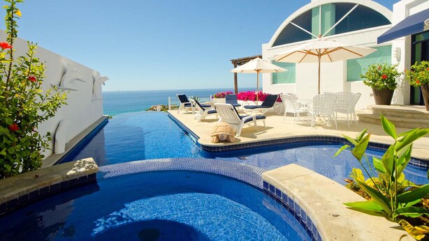 Un espace aménagé au bord de la piscine, avec des chaises longues et des parasols, une vue dégagée sur la mer et un bâtiment moderne doté d’une façade en verre incurvé.