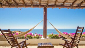 Un hamac tendu entre deux chaises en bois, donnant sur une plage avec une vue dégagée sur la mer et des fleurs roses au premier plan.