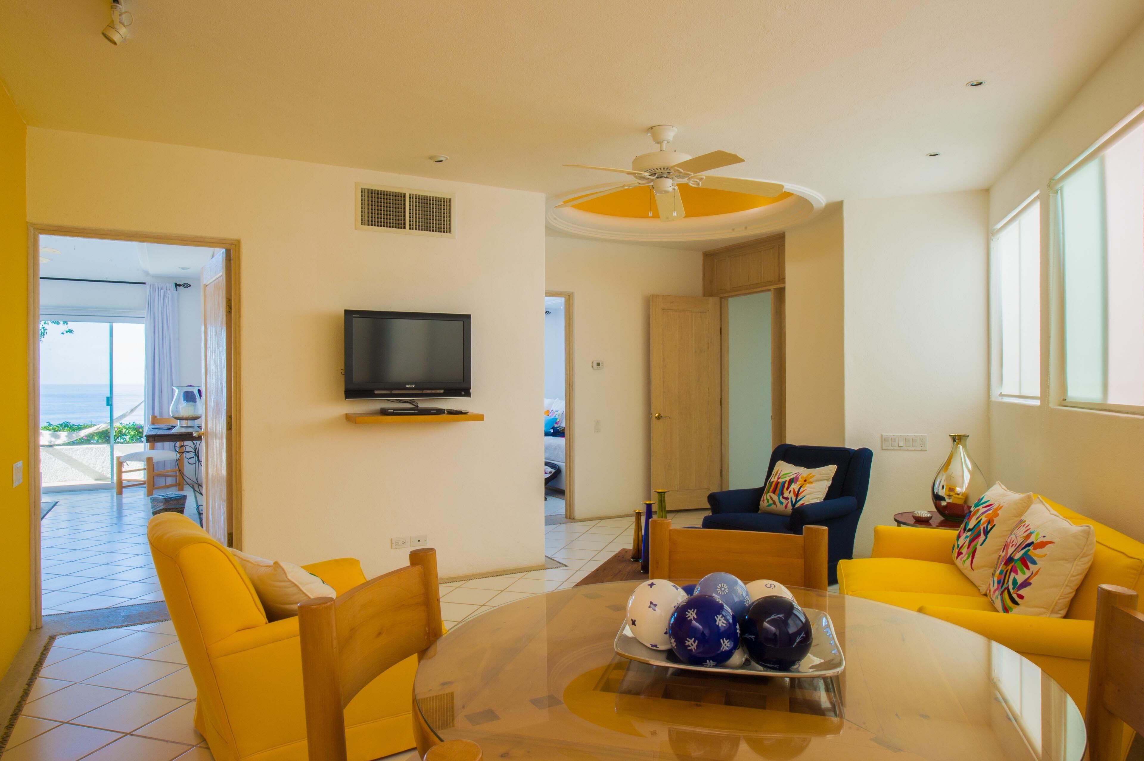 A living room with a TV, yellow sofas, a glass coffee table with sports balls, and a ceiling fan.