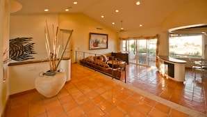 A spacious living area with a large sofa, a coffee table, and a decorative vase. There is a painting on the wall and a window with a view of the outdoors. - Pedregal Oceanview Villa w/ Pool, Hot Tub & Lush Landscaping (Cabo San Lucas)