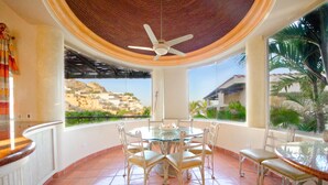A round dining table with six chairs, a ceiling fan, and large windows offering a view of a hillside. - Pedregal Oceanview Villa w/ Pool, Hot Tub & Lush Landscaping (Cabo San Lucas)