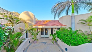 A villa with a tiled roof, a central door, and a surrounding garden with palm trees and flowering plants. - Pedregal Oceanview Villa w/ Pool, Hot Tub & Lush Landscaping (Cabo San Lucas)