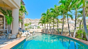 A swimming pool with clear blue water, surrounded by palm trees and rocks, with buildings in the background. - Pedregal Oceanview Villa w/ Pool, Hot Tub & Lush Landscaping (Cabo San Lucas)