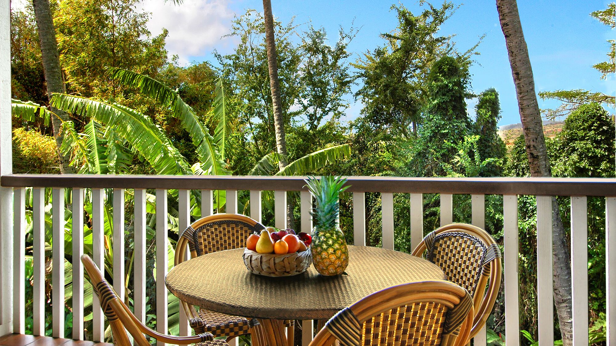Una terraza de madera con mobiliario de mimbre y una mesa dispuesta con frutas y una piña.