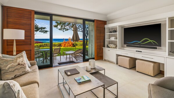 A modern living room with a large TV, comfortable seating, and a sliding glass door leading to a balcony with a view of the ocean.
