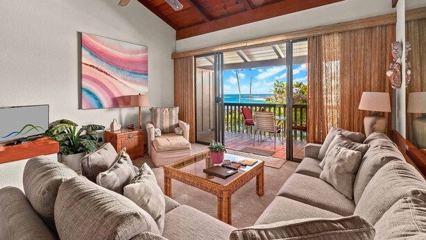 A spacious living room with a large sofa, a coffee table, and a TV. There is a balcony with a view of the ocean.