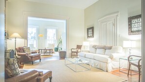 A spacious living room with a large window, a white sofa, a brown leather armchair, and a glass coffee table.