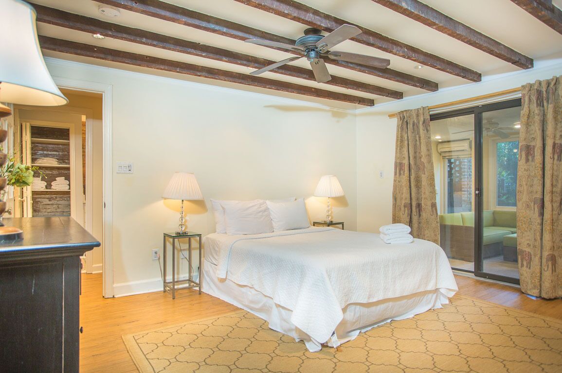 Une chambre à coucher avec un lit, deux lampes de chevet, un ventilateur de plafond et une vue sur l’extérieur, accessible par une porte vitrée.