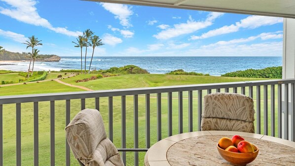 A balcony with a view of the ocean, a grassy area, and a table with a bowl of fruit. - Poipu Sands at Poipu Kai #523 By Parrish Kauai - oceanfront views & steps to bea (Koloa)