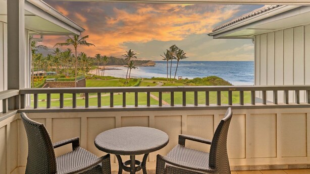 A balcony with wicker chairs and a small round table, overlooking a beach with palm trees and a sunset view.