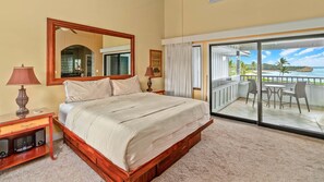 A bedroom with a large bed, a wooden nightstand, a mirror, and a view of a balcony with outdoor furniture. - Poipu Sands at Poipu Kai #523 By Parrish Kauai - oceanfront views & steps to bea (Koloa)