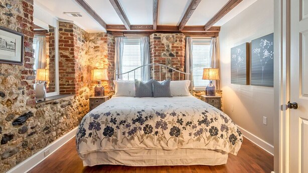 Quarto com parede de pedra, vigas de madeira, cama com roupa de cama floral, duas luminárias de cabeceira e um quadro na parede.
