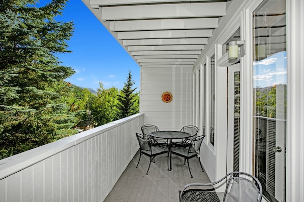 A covered balcony with a table and chairs, a glass door, and a view of trees and mountains.
