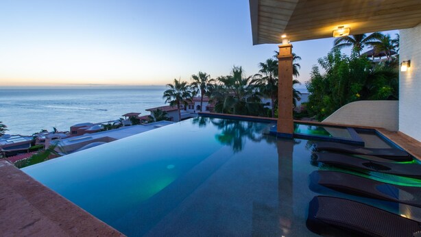 Ein Infinity-Pool mit Blick auf den Ozean, Palmen und eine Holzhütte mit Beleuchtung.
