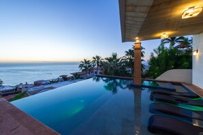 Une piscine à débordement offrant une vue sur l’océan, des palmiers et une pergola en bois illuminée.