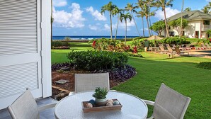 Un patio con una mesa redonda y dos sillas, con vista a un césped cuidado con plantas tropicales y vistas al océano.
