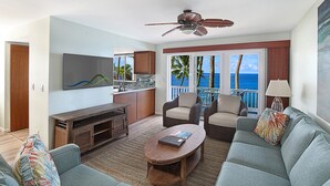 A modern living room with a flat-screen TV, a wooden entertainment unit, a round coffee table, and a sofa with colorful cushions. There is a balcony with a view of the ocean and palm trees. - Poipu Palms #203 By Parrish Kauai: pristine, updated oceanfront gem w/ sunsets & (Koloa)