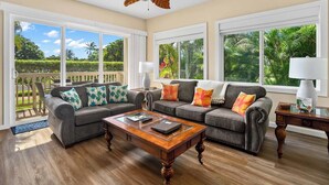 A spacious living room with two grey sofas, a wooden coffee table, and large windows offering a view of a lush garden.