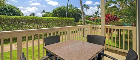 An outdoor patio with a table and chairs, surrounded by a white railing, overlooking a garden with various plants and trees.