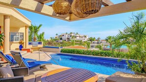 Zona junto a la piscina con sillones de descanso y una mesa, con vista despejada a la piscina y un edificio al fondo.