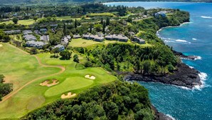 Vista aérea de un complejo turístico costero con varios edificios, un campo de golf y vistas del océano y montañas al fondo.