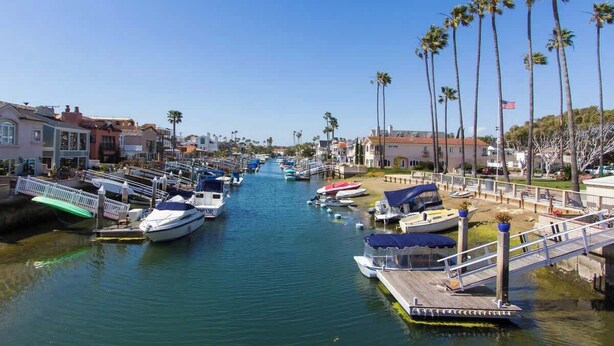 Un port de plaisance où des bateaux sont amarrés le long d’un canal, entouré d’immeubles résidentiels et de palmiers.