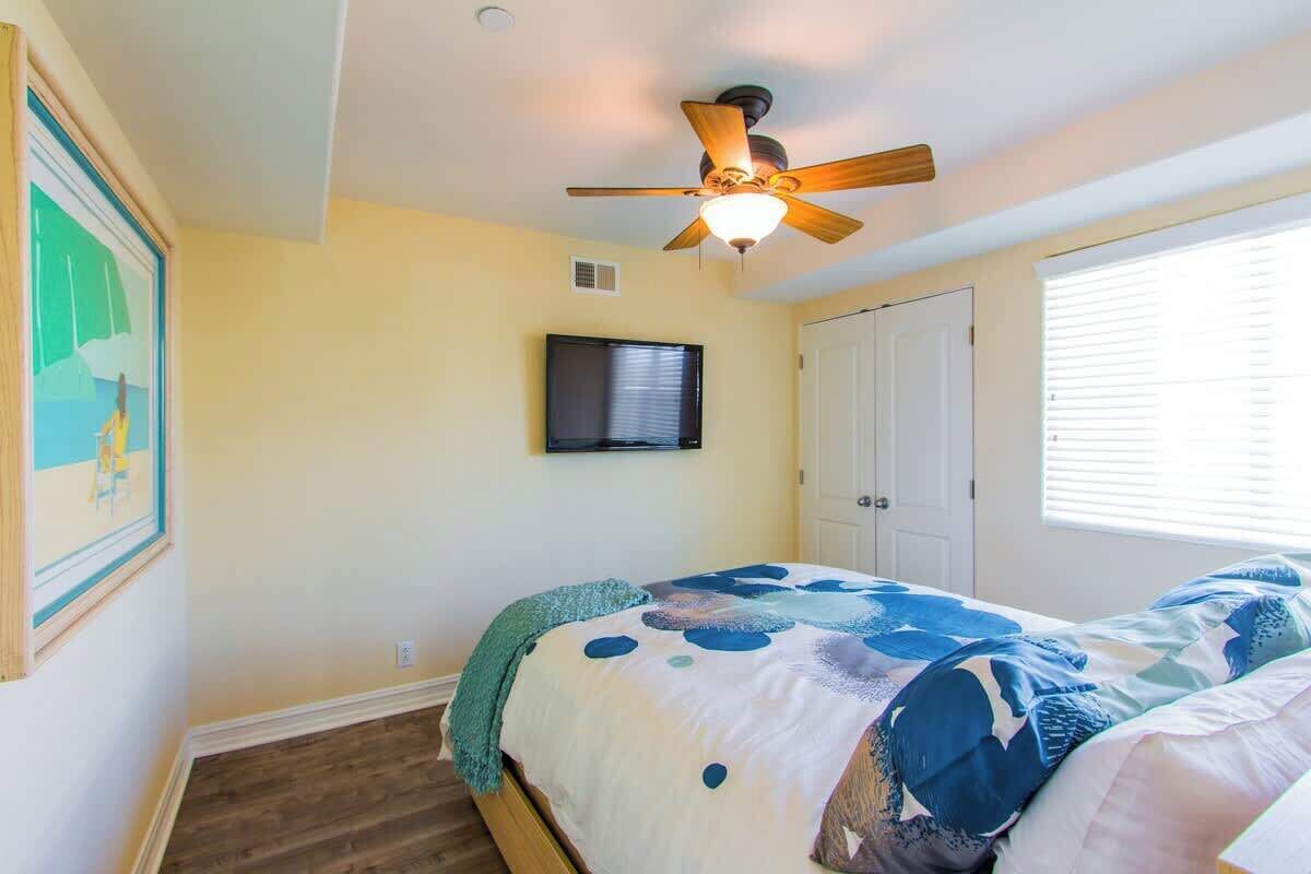 A bedroom with a bed, a ceiling fan, a mounted TV, and a window with blinds.