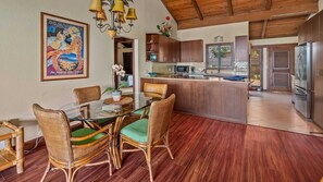 A dining area with a glass table and wicker chairs, a kitchen with wooden cabinets, and a wooden ceiling.