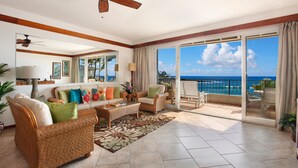 A spacious living room with a balcony overlooking the ocean, featuring wicker furniture, a large window, and a ceiling fan.