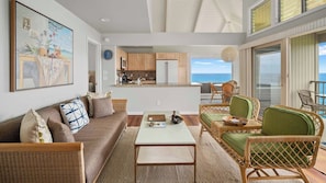 Uma sala de estar com sofá, mesa de centro e cadeiras de vime, uma cozinha com armários de madeira e uma varanda com vista para o mar.