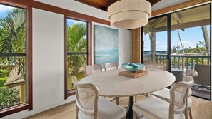 A round dining table with chairs, a hanging light fixture, and large windows with views of palm trees and the ocean.