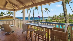 A covered outdoor seating area with wicker furniture, a table, and a view of the ocean and palm trees.