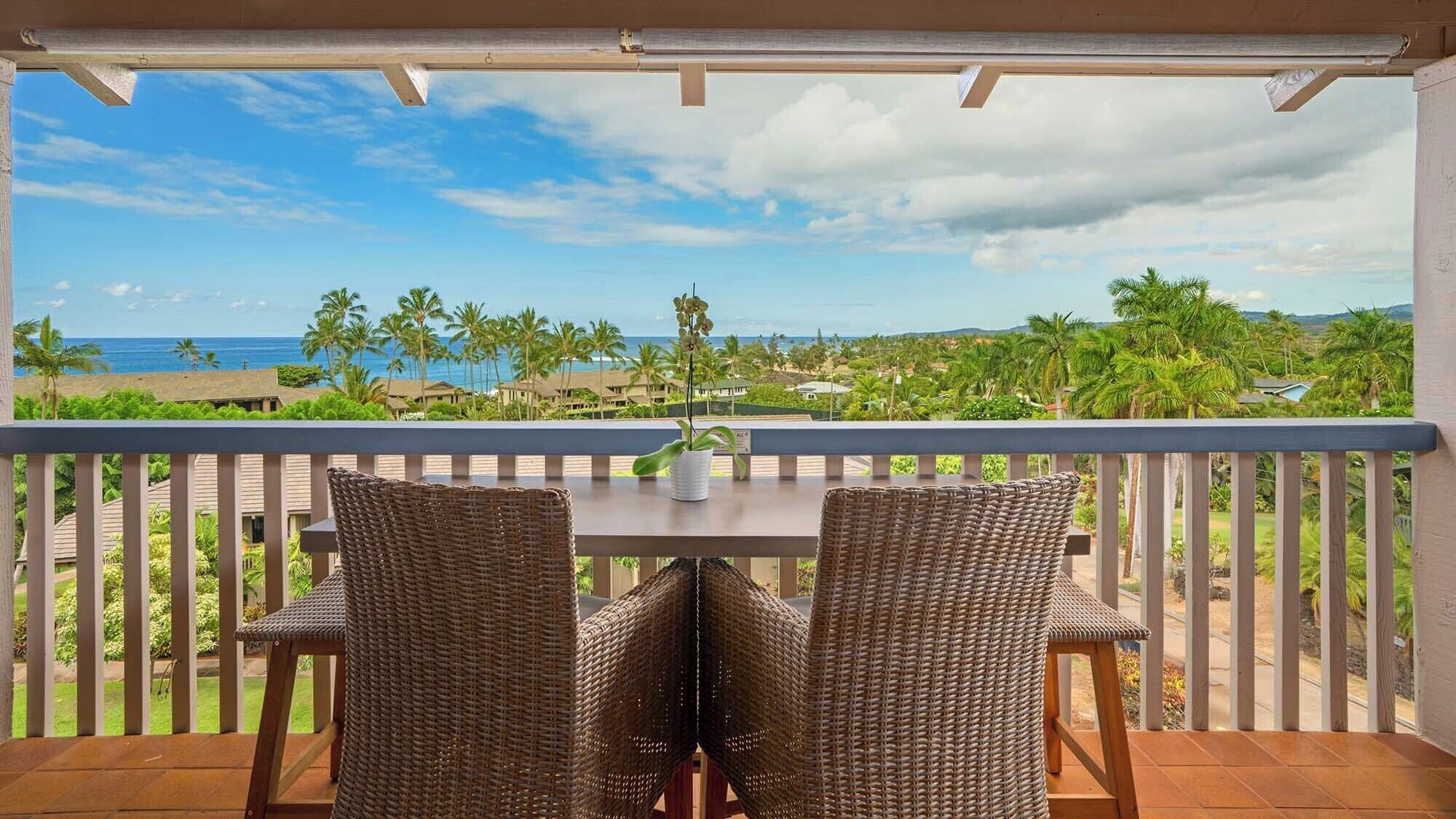 A balcony with wicker chairs and a table, overlooking a beach with palm trees and a view of the ocean.