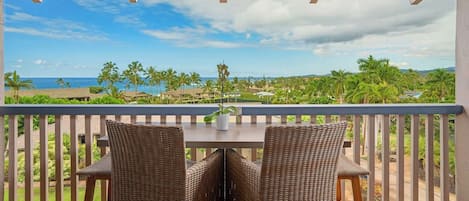 A balcony with wicker chairs and a table, overlooking a beach with palm trees and a view of the ocean.