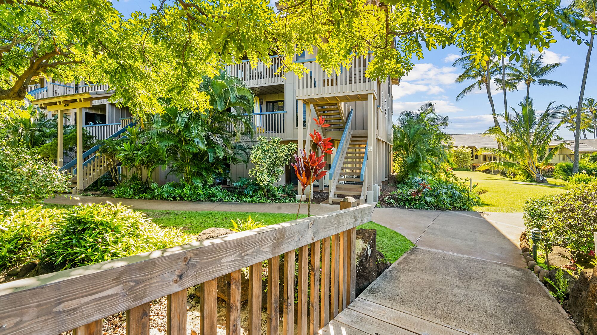 A wooden deck leading to a building with a balcony, surrounded by lush greenery and palm trees.