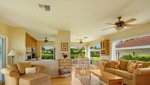 A spacious living room with a sofa, a coffee table, and a dining area with chairs. There is a kitchen area with cabinets and a window overlooking a garden.
