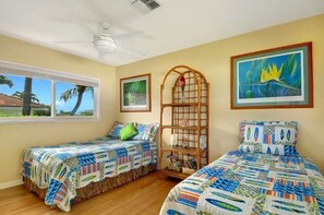 A bedroom with two beds, a window with a view of palm trees, a ceiling fan, and framed artwork on the walls.