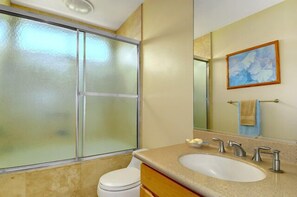 A bathroom with a glass shower enclosure, a sink with two faucets, a mirror, and a framed artwork on the wall.
