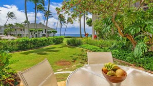 A tropical garden with a view of the ocean, a dining table with a bowl of fruit, and a pool area.