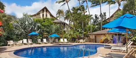 A swimming pool surrounded by lounge chairs and umbrellas, with palm trees and a building in the background.