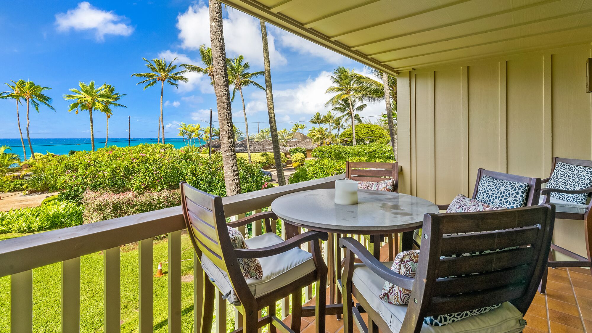 A covered outdoor seating area with a table and chairs, overlooking a tropical landscape with palm trees and a view of the ocean.