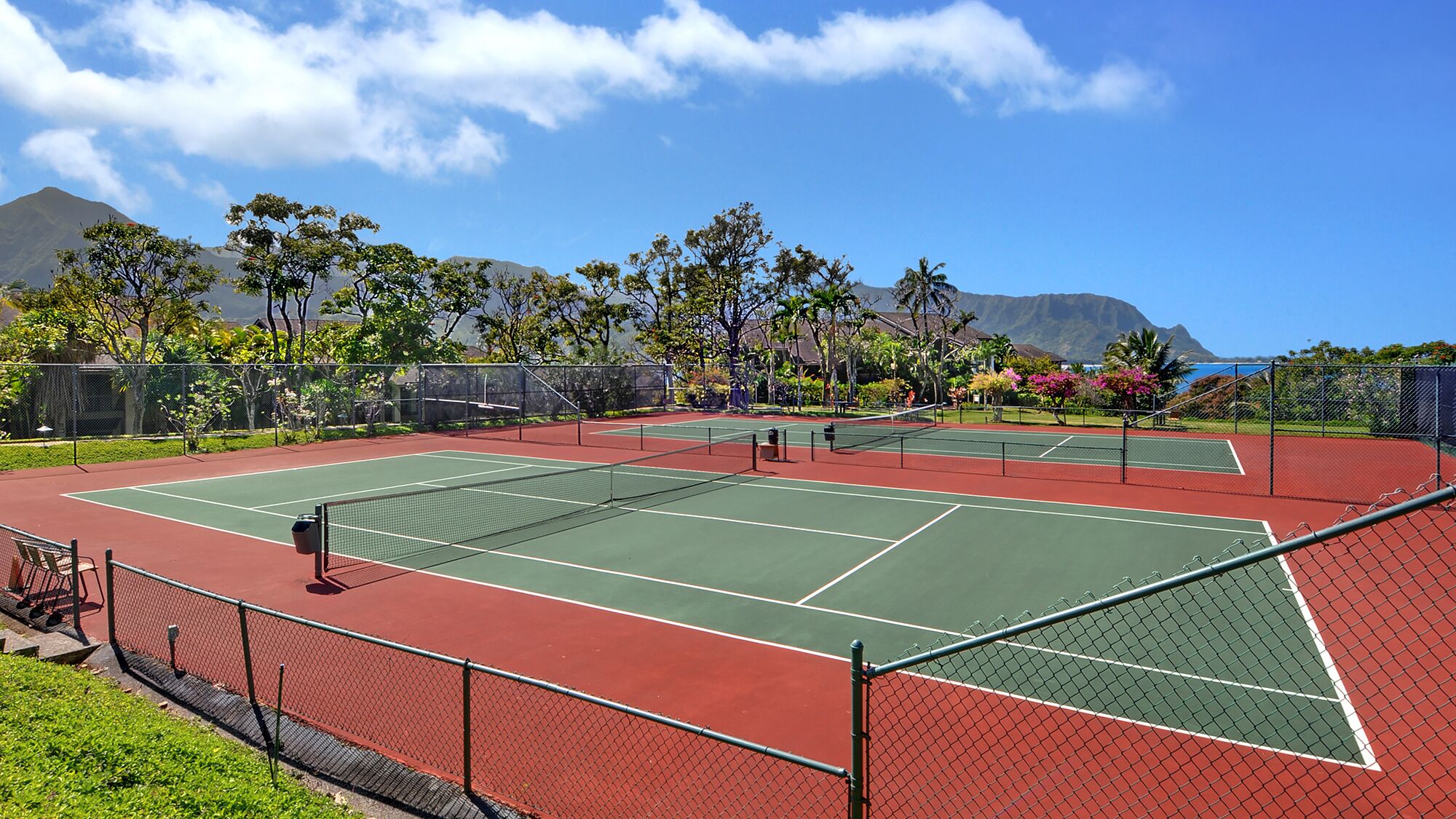 Una cancha de tenis con una red, rodeada por una cerca, y una montaña al fondo.