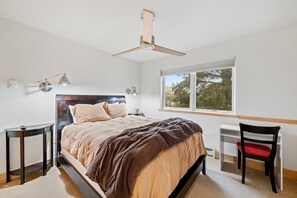 Ein Schlafzimmer mit einem Bett, einem Schreibtisch, einem Stuhl, einem Deckenventilator und einem Fenster mit Blick auf Bäume.